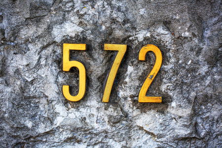 Yellow metal house number 572 mounted on a rough, weathered concrete wall. The bold, golden digits...
