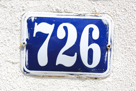 Close-up of vintage enamel house number plaque 726 in white on a blue background. The retro metal...