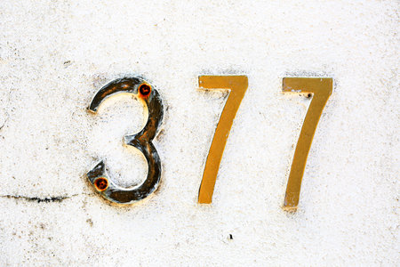 Close-up of weathered metallic house number 377 on a white textured wall. Golden numerals show age...