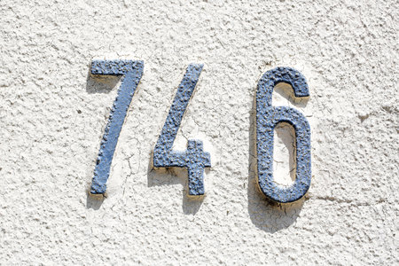 Weathered blue house number 746 mounted on a textured white wall. The painted digits show signs of...