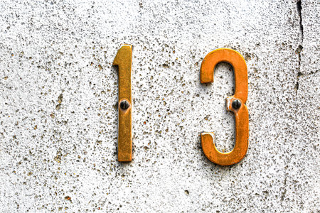 Close-up of golden metal house number 13 mounted on a rough, white stucco wall. The aged digits and...