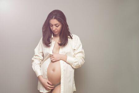 Young pregnant woman in white shirt holding her hands on pregnant belly on gray backgroundの写真素材