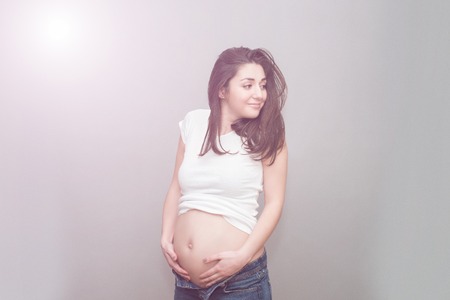 caucasian pregnant girl in unbuttoned jeans and a white t-shirt holding on to naked belly in the studio on a gray backgroundの写真素材
