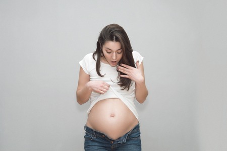 caucasian pregnant girl in unbuttoned jeans and a white t-shirt holding on to naked belly in the studio on a gray backgroundの写真素材
