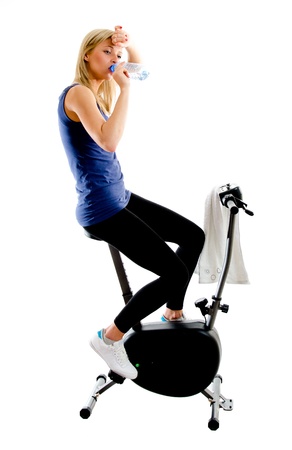Tired girl drinking mineral water during exercising on stationary training bike - isolated on white backgroundの写真素材