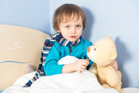 Little sick baby boy checking teddy bear's body temperature in bedの写真素材