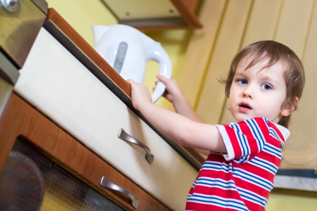 Little baby boy is reaching eletrical kettle with hot water - danger in kitchenの写真素材
