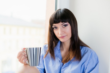 A beautiful brunette in blue shirt drinking he first morning cup of coffee or tea while sitting by the windowの写真素材