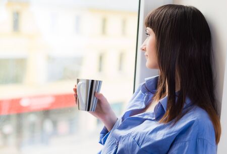 Woman in blue shirt with a cup of tea looking through the windowの写真素材