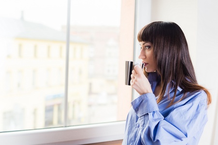 Woman standing by the window drinking coffee or tea from a cupの写真素材
