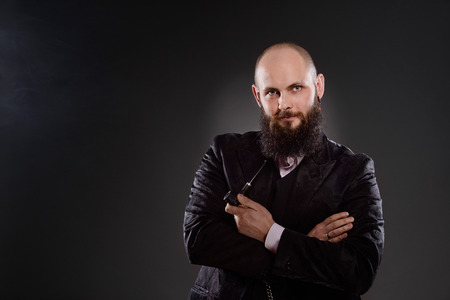 Bald bearded man holding a pipe in crossed hands.He is dressed in a black jacket and vest. Looking at camera. Photo taken in the studio on a gray backgroundの写真素材