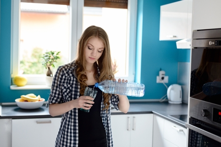 Young blonde woman in kitchen woman pouring a glass of water. Standing in a bright modern kitchen. Dressed in a plaid shirt with short sleeveの写真素材