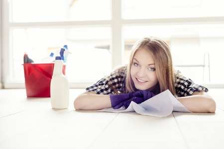 Beautiful young woman resting during cleaning of the apartment. Lying on her stomach and holding a cloth. Looking at camera, smiling. Next to her a bucket of cleaning utensilsの写真素材