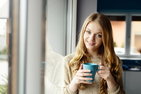Beautiful smiling blond woman drinking coffee by the window. She looks at the cameraの写真素材