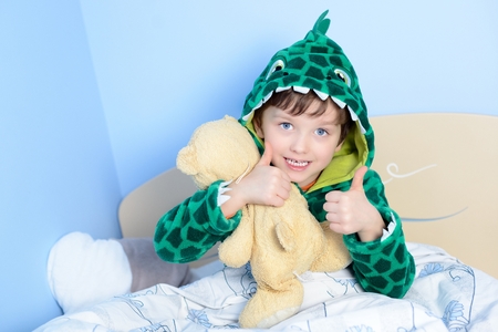 Portrait of little boy gesturing thumbs up with teddy bear in bed.  A child dressed in a funny bathrobeの写真素材