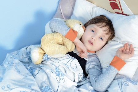 Portrait of a sick boy hugging a teddy bear lying in a bedの写真素材