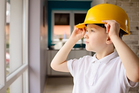 6 year old boy posing by a window with a yellow helmet on his head. Adjusting helmet on his head