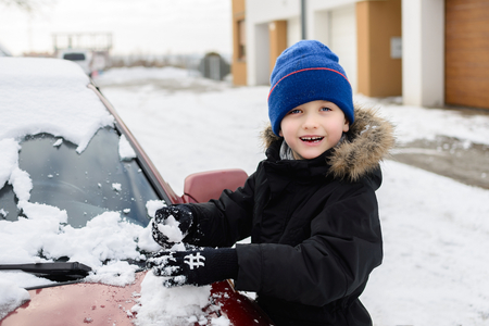 Child playing with snow outside. Taking the snow from the snow-covered car Dressed in a black jacket and blue capの写真素材
