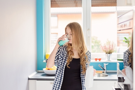 Young blond woman drinking milk from the glass. Standing in the kitchen. Dressed in a plaid shirtの写真素材