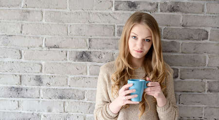 A beautiful young blonde woman drinking coffee from a cup. She stands near a brick wallの写真素材