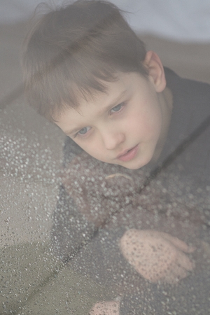 Child is hugging his teddy bear. Sitting behind wet from rain window glass. Rainy Day. Loneliness and waiting conceptの写真素材