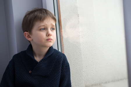 Sad 7 years boy child looking out the window. Rainy day