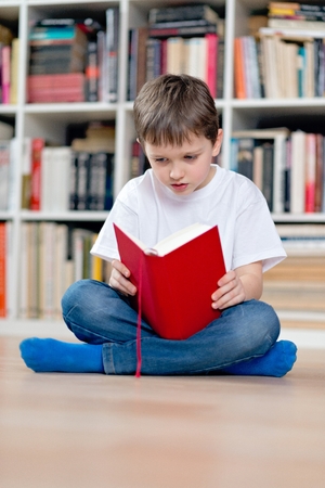 Little boy child reading a red book. He sits cross-legged on the floor. Dressed in a white t shirt and blue jeansの写真素材