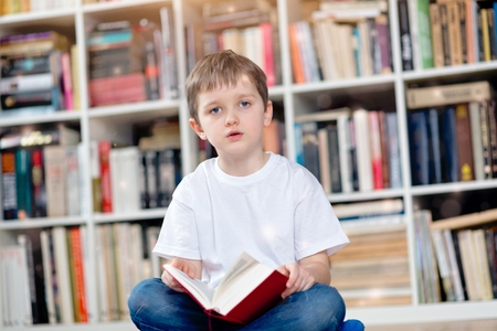Little boy with red book in the library. Looking at camera. He sits cross-legged on the floor. Dressed in a white t shirt and blue jeansの写真素材