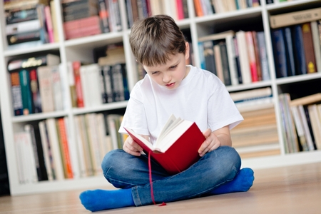 Child reading a red book in the library. He sits cross-legged on the floor. Dressed in a white t shirt and blue jeansの写真素材