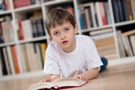 Boy lies on his stomach and reading a book in the library. Dressed in a white t shirt and blue jeans. Looking at camera.の写真素材