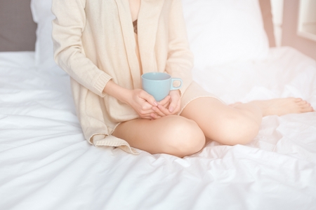 Young woman drinking her coffee while sitting on bed. Lazy sunny morning in bed. Dressed in beige sweater.の写真素材