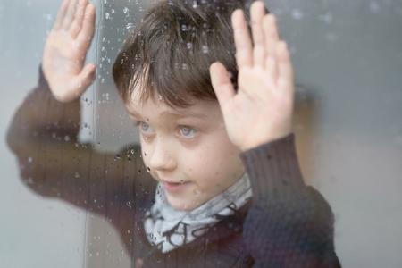 Young 7 years old boy standing beside the window with raindrop on the glass after rain. Rainy Dayの写真素材