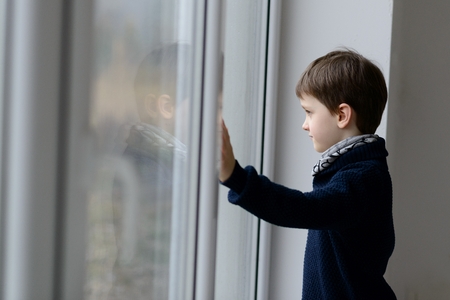 Thoughtful 7 year old boy standing by the window. Rainy day.の写真素材