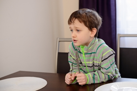 Little 7 years old boy holding fork and knife and waiting for dinnerの写真素材