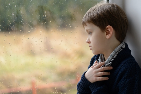 Sad thoughtful little boy looking through the window. Copy space for text on glass. Rainy dayの写真素材