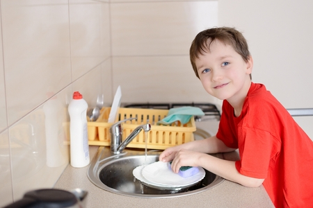 smiling 7 year old boy washes dishes in the kitchen. Dressed in a red t-shirtの写真素材