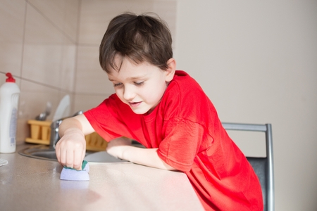 Satisfied 7 year old boy cleans cabinets in the kitchen. Dressed in a red t-shirtの写真素材