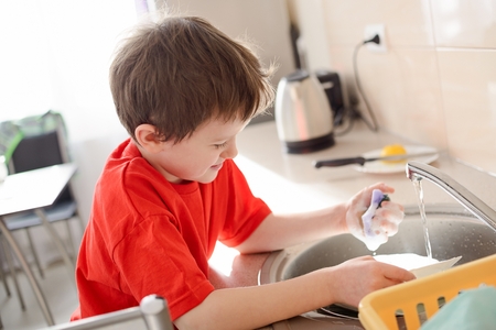 boy washes dishes in the kitchen. Dressed in a red t-shirtの写真素材