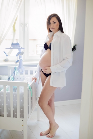 Beautiful brunette 8 months pregnant woman posing in home. Dressed in white shirt and black lingerieの写真素材