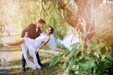 The groom is going to kiss the bride leaning her and holding his hand under her knee. Wedding session in the park near lakeの写真素材