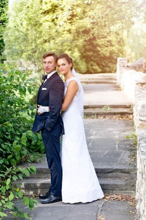 The bride standing behind groom and hugs him. Groom holds his hands in his pockets. Wedding session in the parkの写真素材