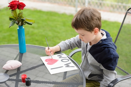 Little 7 year old boy paints greeting card for Mom on Mother's Day with the inscription "I love you mom". Outdoors. Mother's Dayの写真素材