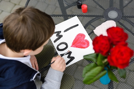 Little 7 year old boy paints greeting card for Mom on Mother's Day with the inscription "I love you mom". Outdoors. Mother's Dayの写真素材