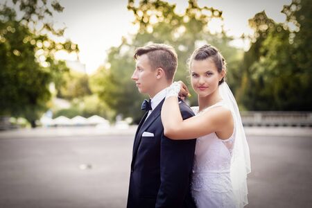 Wedding session in park. Bride standing behind groom and looking at cameraの写真素材