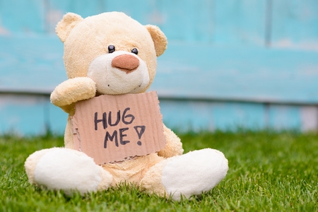 Little old teddy bear sitting on the grass in the garden and holding a piece of cardboard with the information - hug meの写真素材