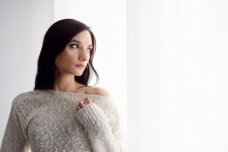 Beautiful young brunette woman in woolen sweater standing near window.の写真素材