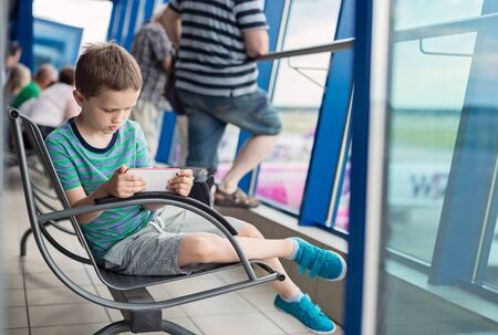 Bored 7 years old boy child waiting for his plane at airport and play games on smartphoneの写真素材
