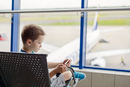Bored 7 years old boy child waiting for his plane at airport and play games on smartphoneの写真素材