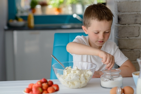 7 year old boy adding sugar to cottage cheese in a bowl. Prepares mini cheesecakes with strawberriesの写真素材