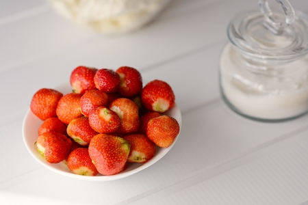 Bowl full of red fresh strawberries. Next to bowl cup with sugar. White wooden table.の写真素材